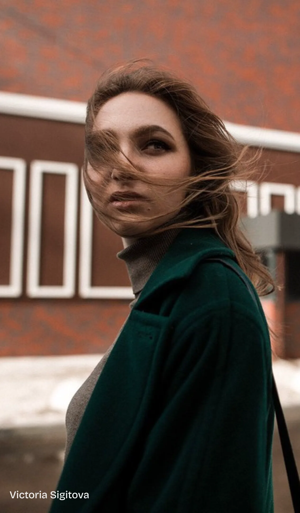 Victoria Sigitova in a dark green coat, hair in the wind, in front of a brick building